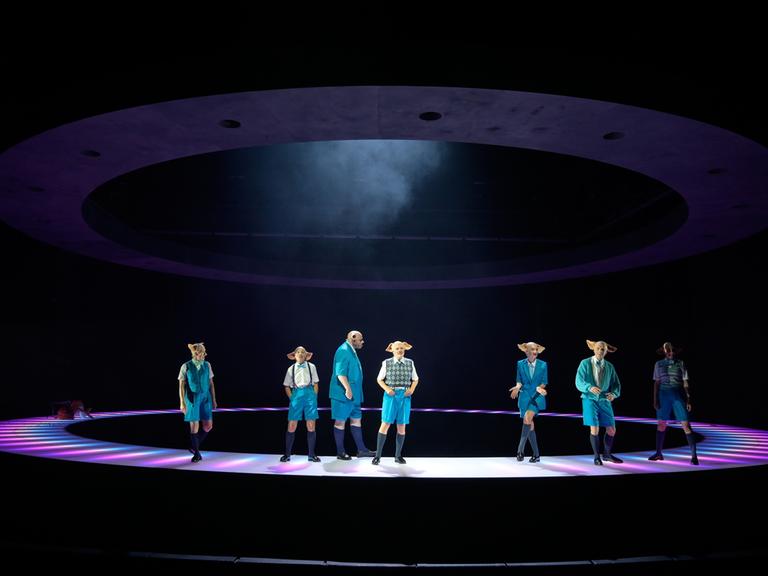 Sieben bunt gekleidete Schauspielerinnen und Schauspieler mit Schweinemasken stehen im Kreis auf der Bühne.

