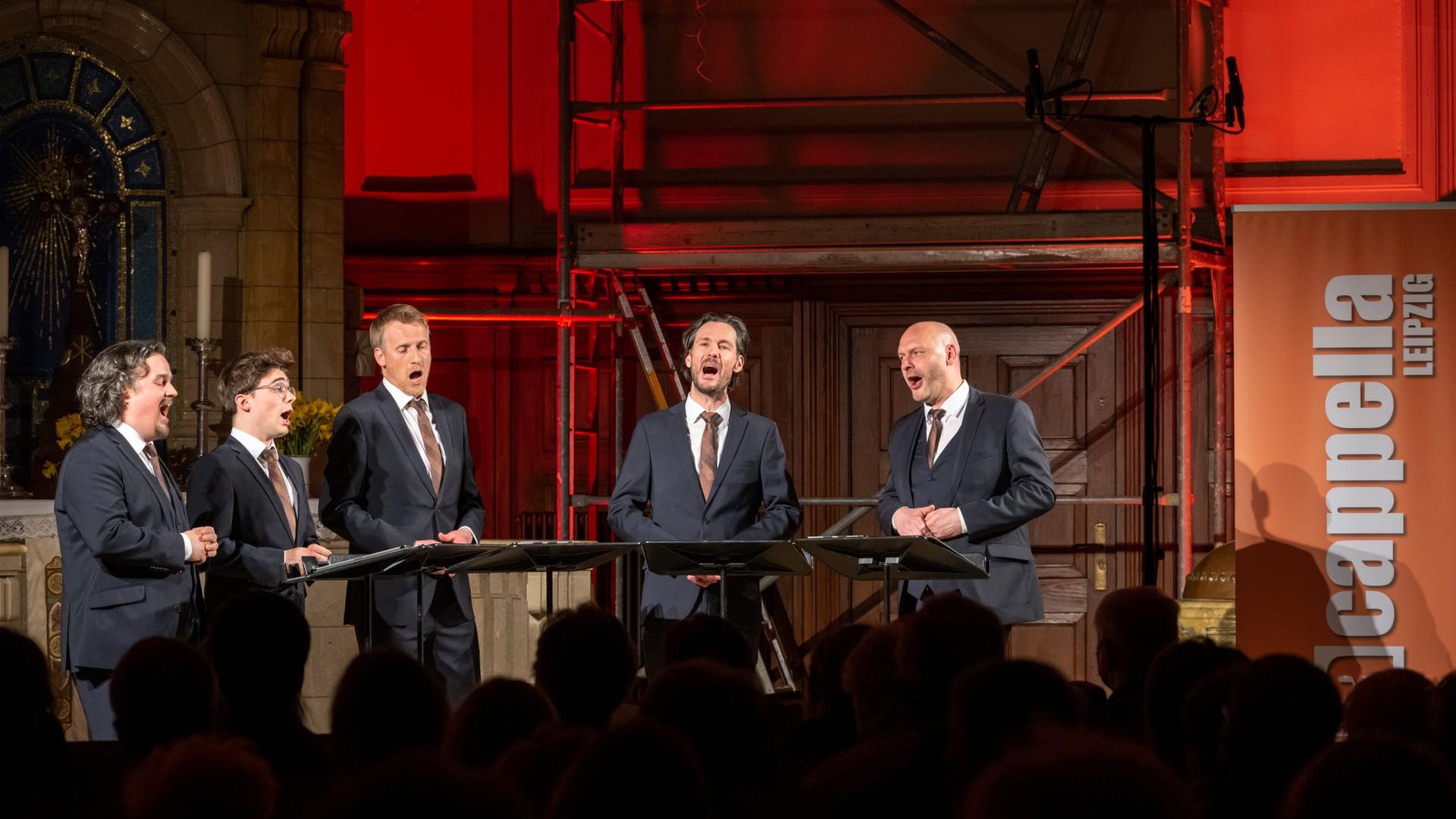 Fünf Männer stehen im Halbkreis im Altarbereich einer Kirche, sie singen und tragen blaue Anzüge. Die Kirchenwände sind rot angestrahlt. Fünf Männer stehen im Halbkreis im Altarbereich einer Kirche, sie singen und tragen blaue Anzüge. Die Kirchenwände sind rot angestrahlt.