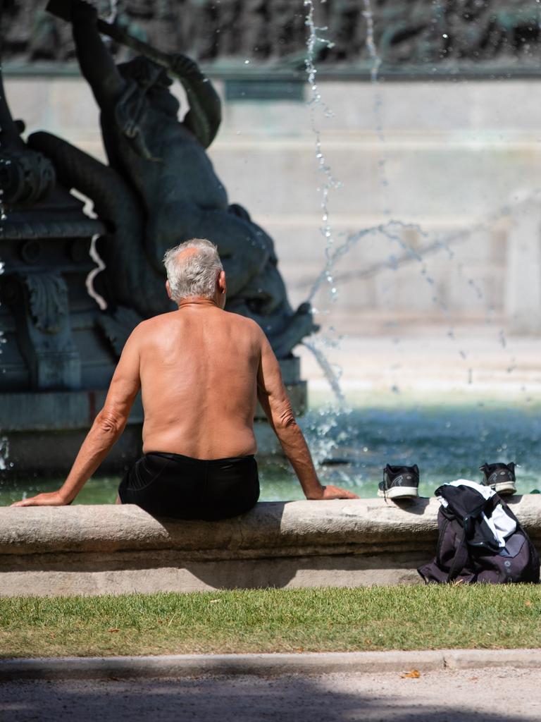 Ein älterer Mann sitzt bei heißen Temperaturen oben ohne auf dem Rand eines Brunnens, seine Kleidung liegt neben ihm. 