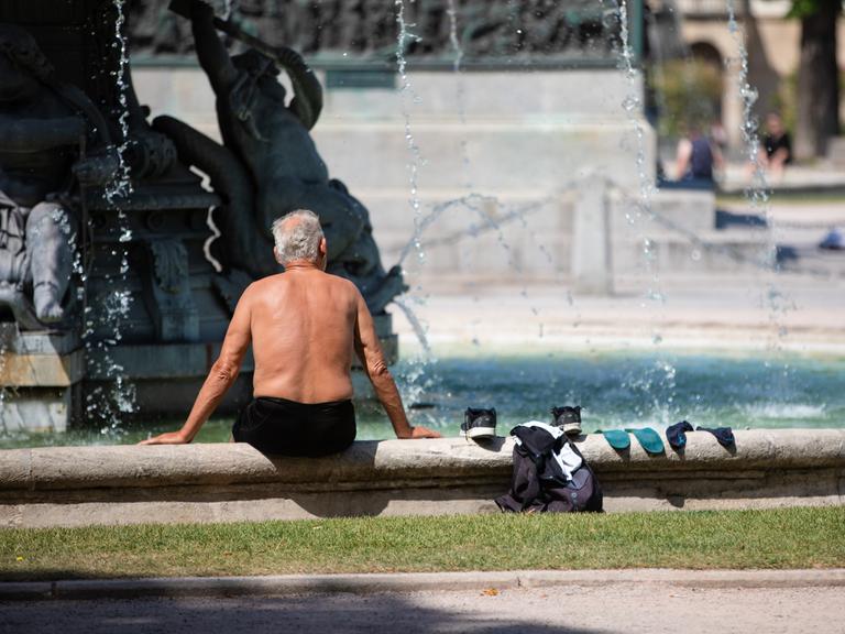 Ein älterer Mann sitzt bei heißen Temperaturen oben ohne auf dem Rand eines Brunnens, seine Kleidung liegt neben ihm. 
