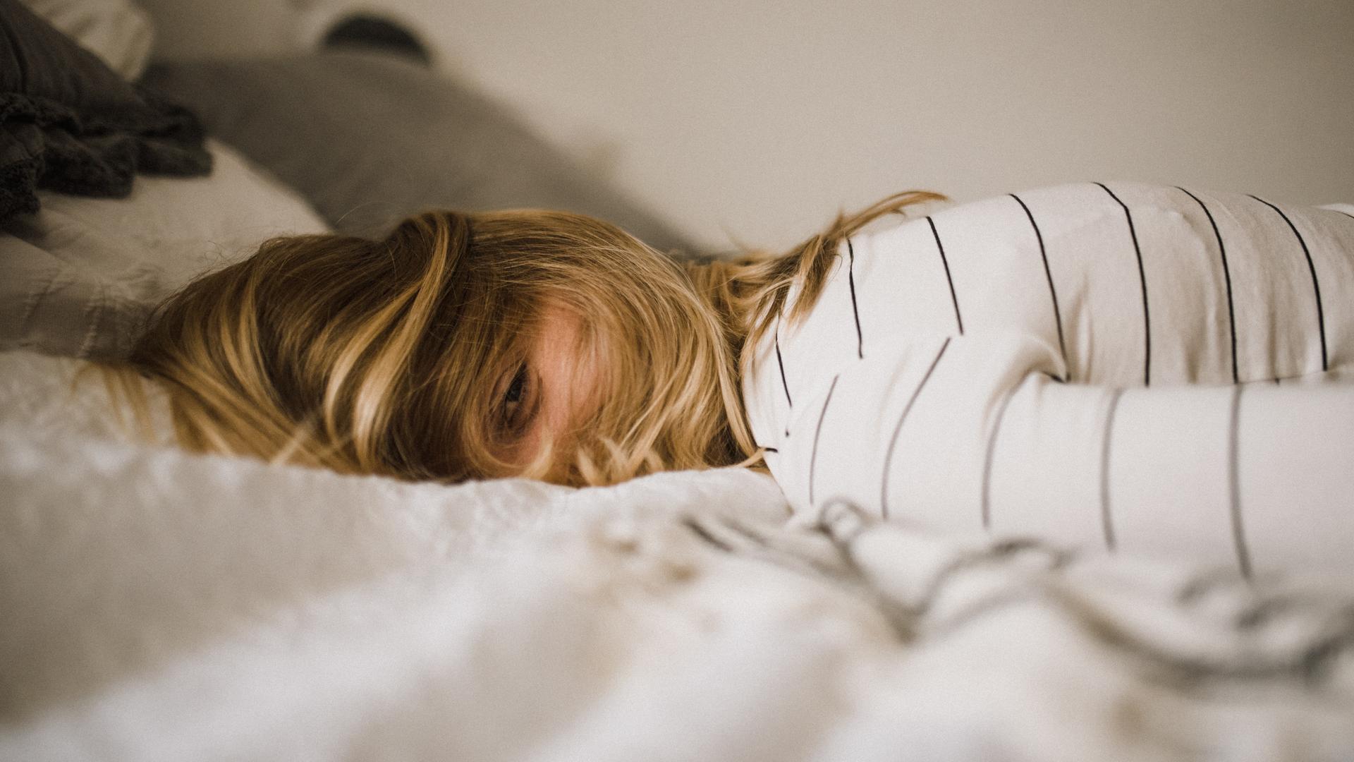 Eine Person mit langen blonden Haaren über dem Gesicht und halb geöffneten Augen liegt auf einem Bett. Eine Person mit langen blonden Haaren über dem Gesicht und halb geöffneten Augen liegt auf einem Bett.