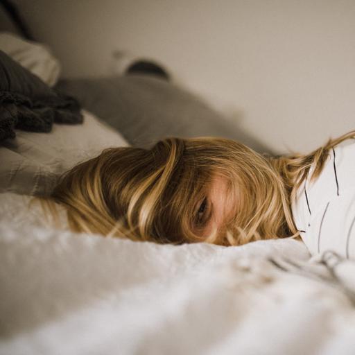 Eine Person mit langen blonden Haaren über dem Gesicht und halb geöffneten Augen liegt auf einem Bett.