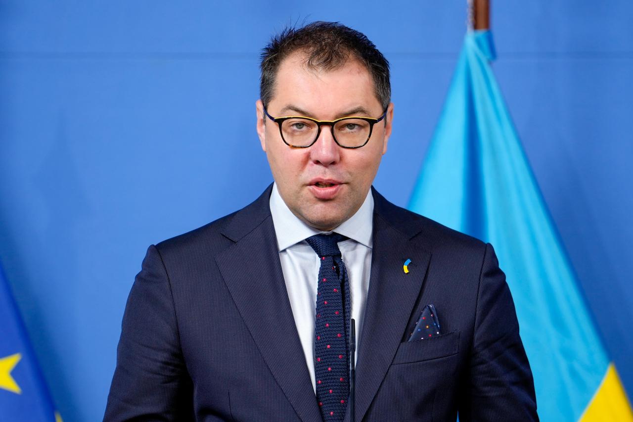 Makeiev im dunklen Anzug steht vor einer blauen Wand, hinter ihm eine ukrainische Flagge.