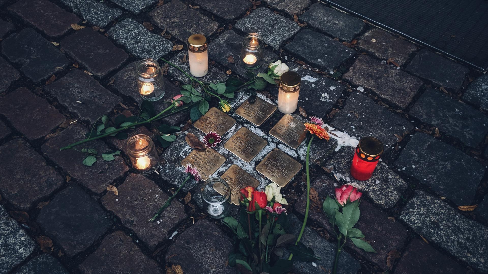 Kerzen und Blumen liegen um eine Ansammlung von sieben Stolpersteinen herum. Kerzen und Blumen liegen um eine Ansammlung von sieben Stolpersteinen herum.