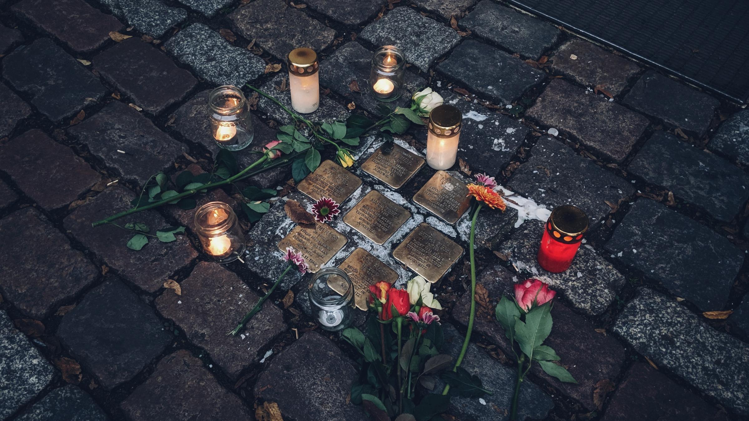 Kerzen und Blumen liegen um eine Ansammlung von sieben Stolpersteinen herum.