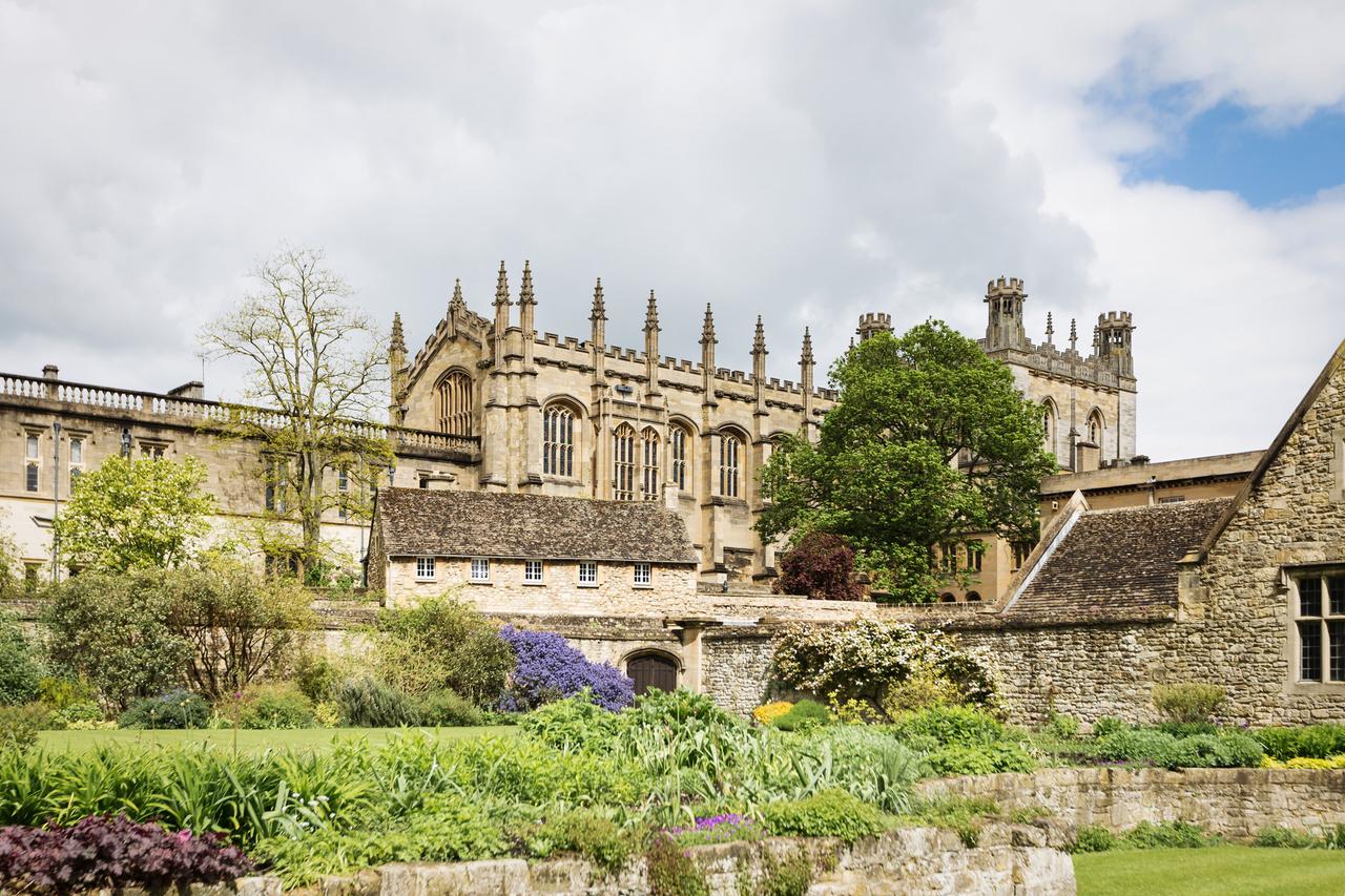 Eine aktuelle Aufnahme zeigt das historische Gebäude des Christ Church College in Oxford inmitten von Blumenrabatten und Bäumen.