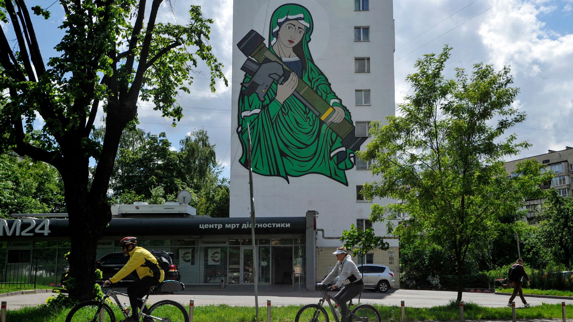 Radfahrer fahren an einem Wandgemälde vorbei, das die heilige Maria mit einer Panzerabwehrlenkwaffe in den Händen zeigt. Radfahrer fahren an einem Wandgemälde vorbei, das die heilige Maria mit einer Panzerabwehrlenkwaffe in den Händen zeigt.