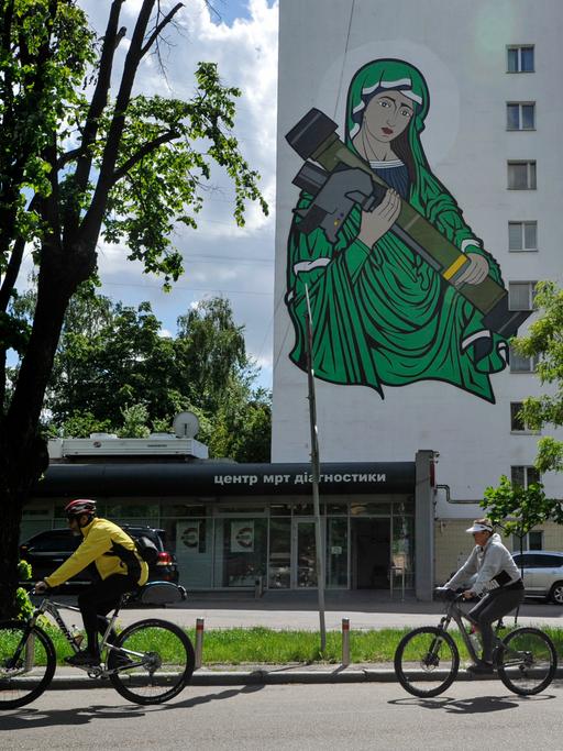 Radfahrer fahren an einem Wandgemälde vorbei, das die heilige Maria mit einer Panzerabwehrlenkwaffe in den Händen zeigt.
