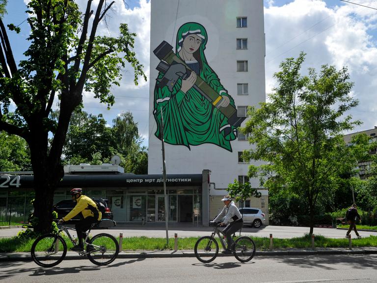 Radfahrer fahren an einem Wandgemälde vorbei, das die heilige Maria mit einer Panzerabwehrlenkwaffe in den Händen zeigt.