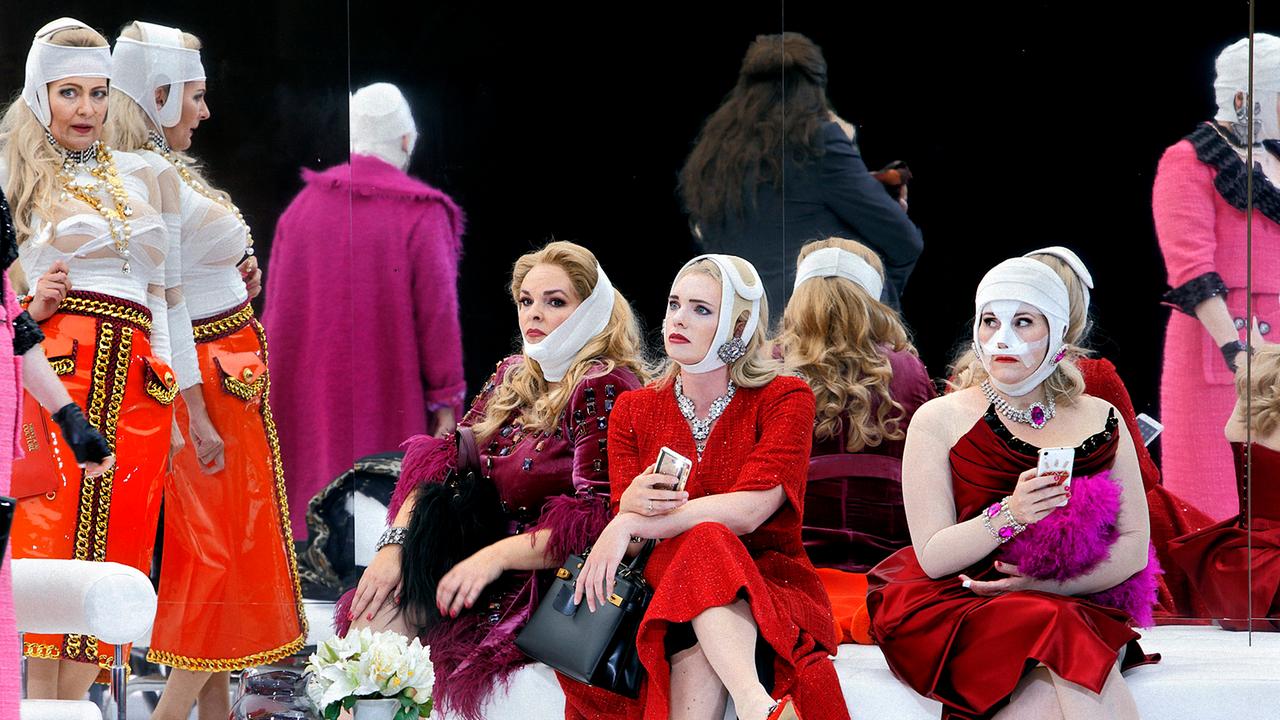 Frauen in Kleidern sitzen und stehen herum. Sie tragen Bandagen von Schönheits-OPs. Manche haben ein Smartphone in der Hand.