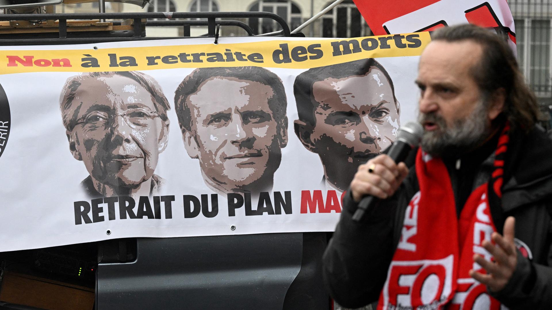 Ein Mann spricht in ein Mikrofon. Er trägt einen Schal mit dem Logo der Gewerkschaft Force Ouvrière. Im Hintergrund sieht man ein Protestbanner mit den Köpfen führender französischer Politiker darauf, unter ihnen ist auch Präsident Macron.