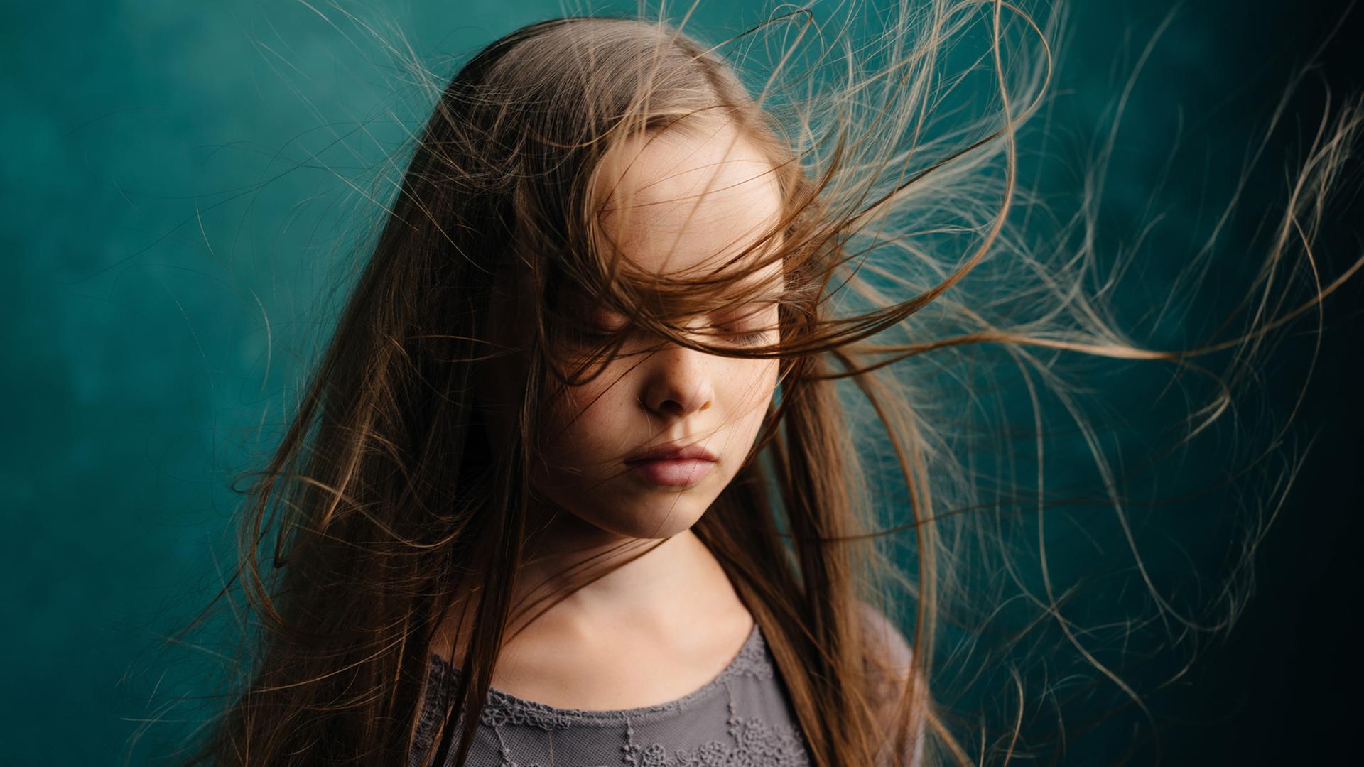 Trauernden Kindern und Jugendlichen beizustehen führt oft auch Erwachsene an emotionale Grenzen. Zu sehen: Ein Mädchen mit geschlossenen Augen und wehenden Haaren. Der Hintergrund ist eine grüne Wand. 