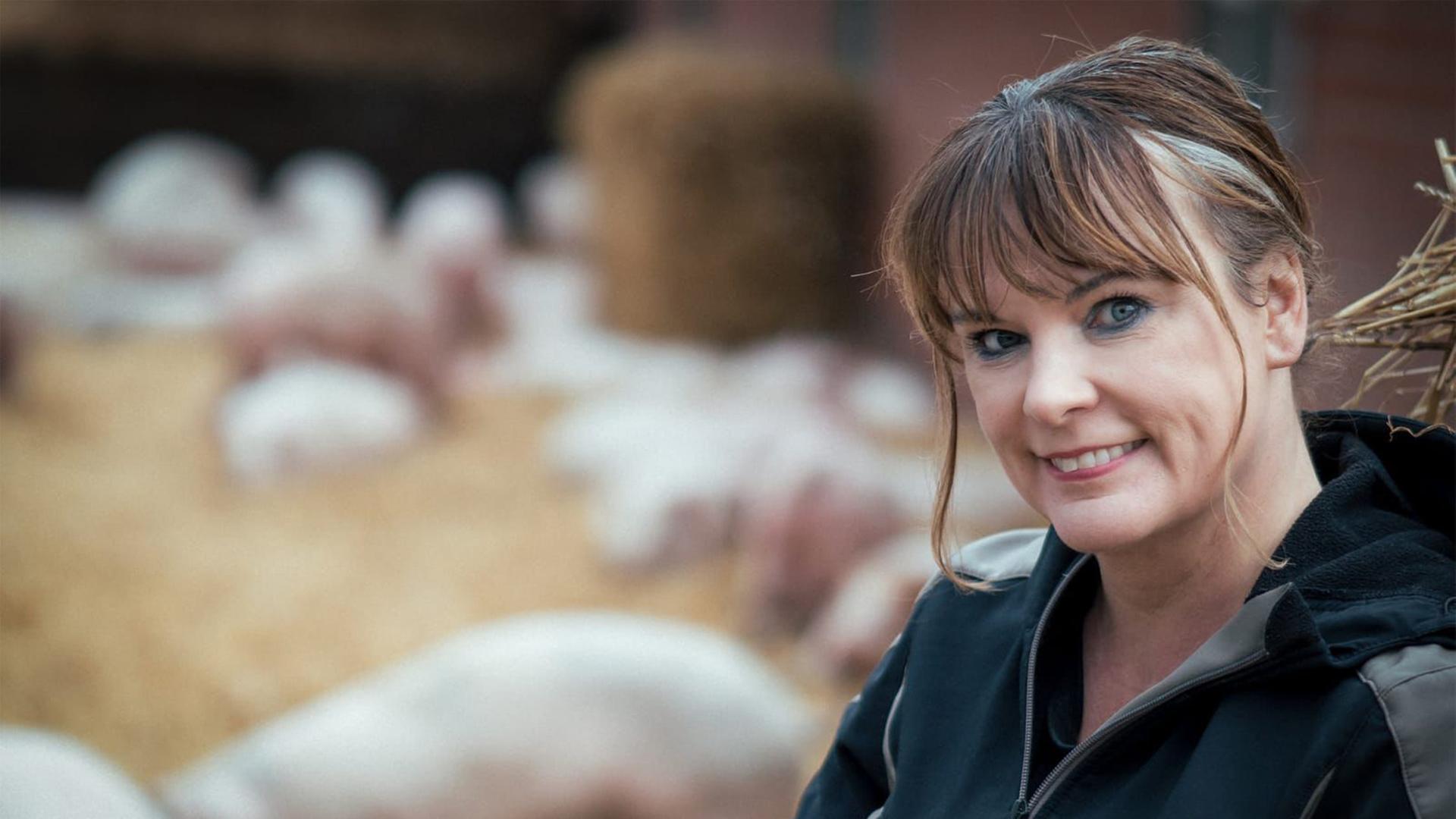 Gabriele Mörixmann trägt eine Wetterjacke und blickt freundlich in die Kamera. Im Hintergrund: Ferkel im Auslauf. Gabriele Mörixmann trägt eine Wetterjacke und blickt freundlich in die Kamera. Im Hintergrund: Ferkel im Auslauf.