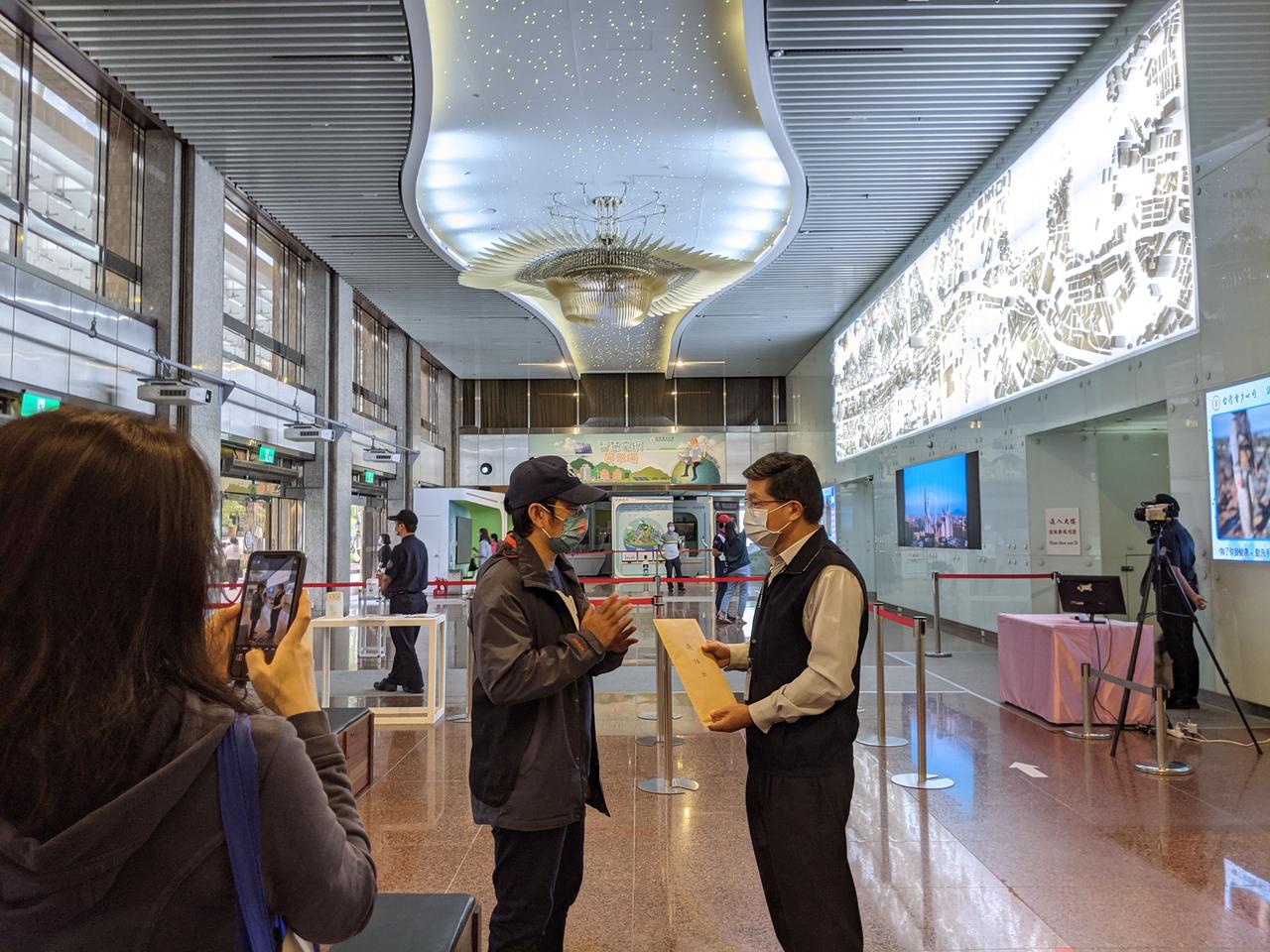 Zwei Männer mit Mund-Nasenschutz stehen sich in einem Foyer gegenüber. Einer hält eine Mappe mit einem Brief in der Hand.