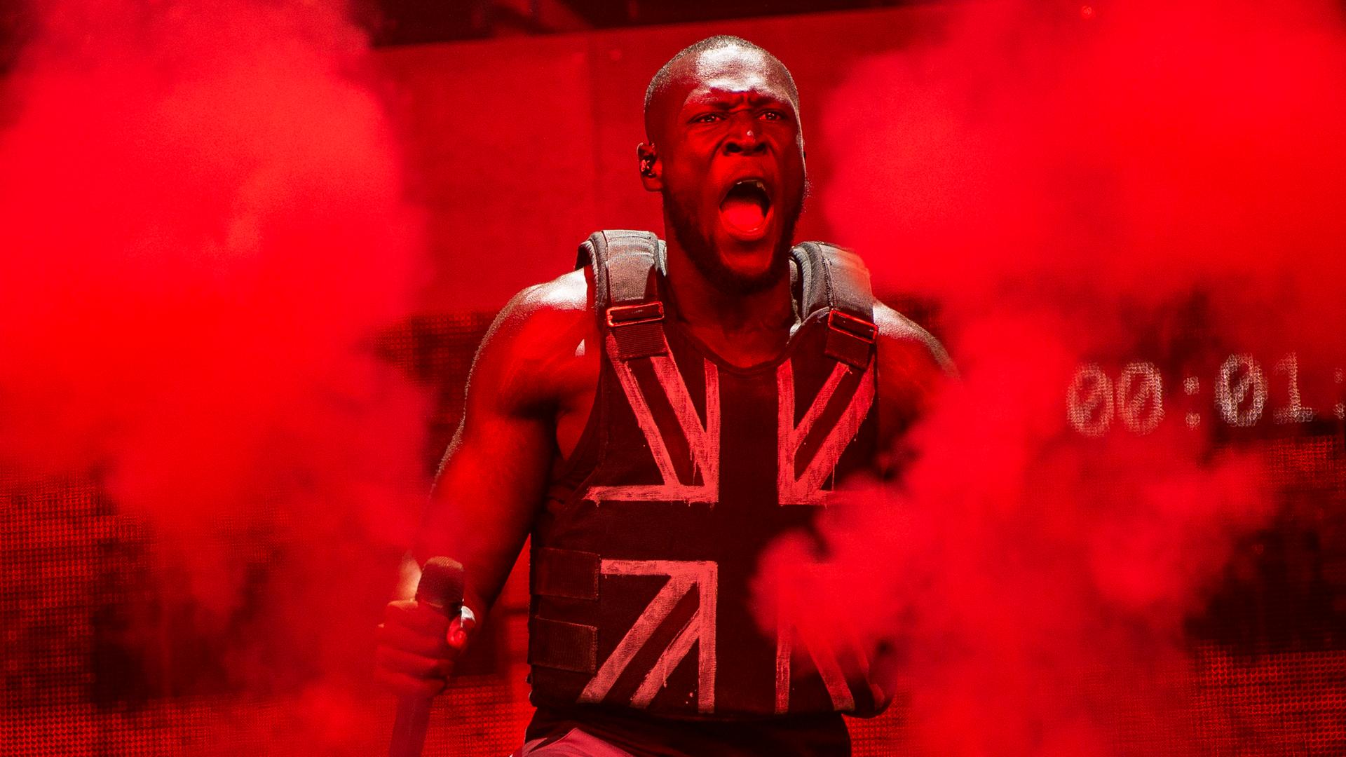 Musiker Stormzy steht auf einer in rotes Licht getauchte Bühne, er hat ein Mikro in der Hand und trägt ein Oberteil im Union-Jack-Look. Musiker Stormzy steht auf einer in rotes Licht getauchte Bühne, er hat ein Mikro in der Hand und trägt ein Oberteil im Union-Jack-Look.