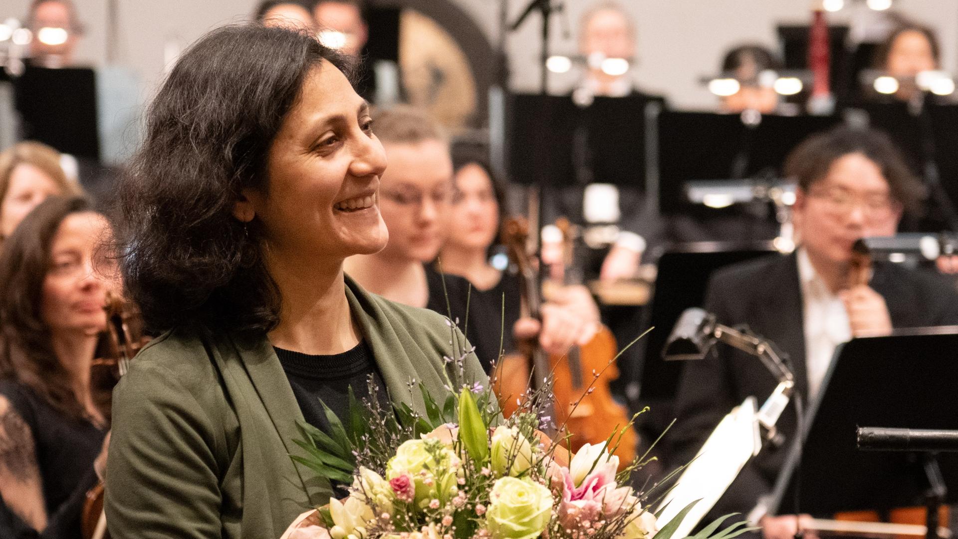 Farzia Fallah nimmt Beifall und Blumenstrauß nach der Uraufführung vor dem Orchester stehend entgegen. 