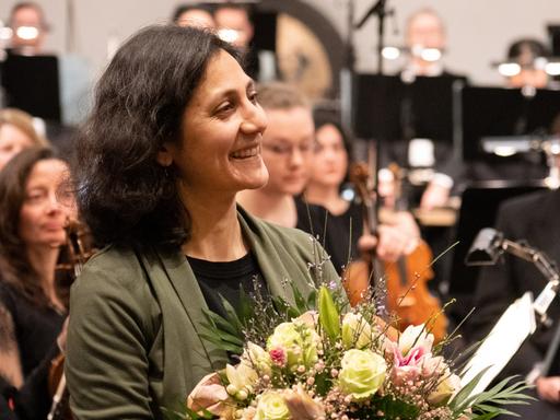 Farzia Fallah nimmt Beifall und Blumenstrauß nach der Uraufführung vor dem Orchester stehend entgegen. 
