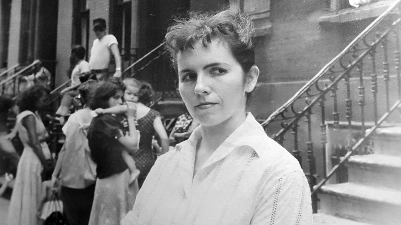 Schwarzweißfoto von Grace Paley als junger Frau in weißer Bluse und mit zusammengebundenem Haar. Hinter ihr vor einem Hausaufgang sind unscharf weitere junge Frauen mit Kindern auf dem Arm zu sehen. Schwarzweißfoto von Grace Paley als junger Frau in weißer Bluse und mit zusammengebundenem Haar. Hinter ihr vor einem Hausaufgang sind unscharf weitere junge Frauen mit Kindern auf dem Arm zu sehen.