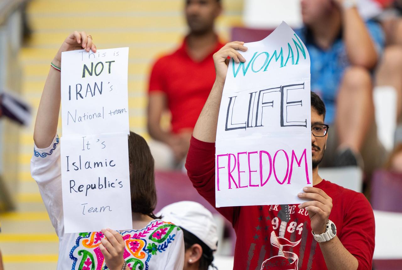 "Das ist nicht die iranische Nationalmannschaft, sondern die der Islamischen Republik", schreibt diese Frau auf einem Plakat. Ein Mann neben ihr hält ein Schild mit "Women – Life – Freedom" in die Luft. "Das ist nicht die iranische Nationalmannschaft, sondern die der Islamischen Republik", schreibt diese Frau auf einem Plakat. Ein Mann neben ihr hält ein Schild mit "Women – Life – Freedom" in die Luft.
