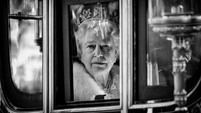 Die Königin ist durch das Fenster ihrer Kutsche zu sehen. Sie schaut direkt in die Kamera. Sie trägt ein Diadem mit Diamanten auf dem Kopf.