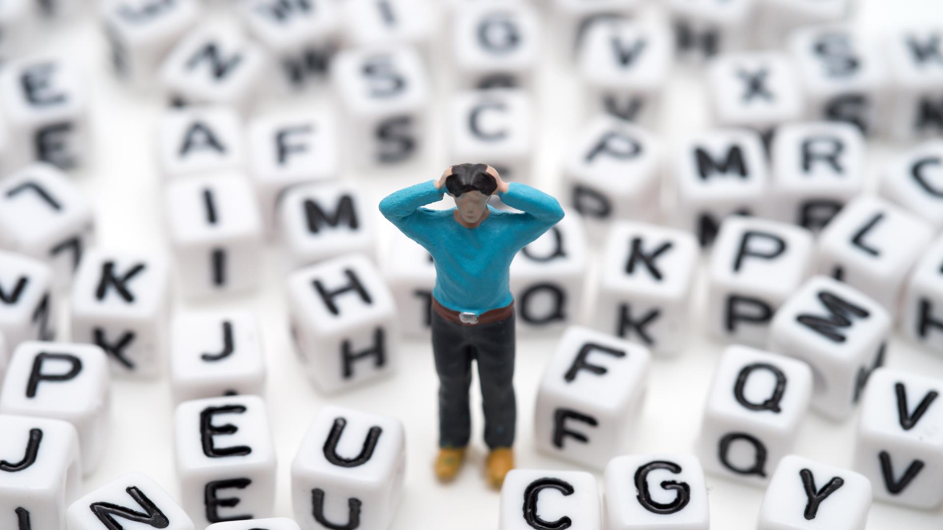 Eine kleine Spielfigur steht, die Hände an den Kopf gehoben, zwischen lauter Buchstabenwürfeln Eine kleine Spielfigur steht, die Hände an den Kopf gehoben, zwischen lauter Buchstabenwürfeln