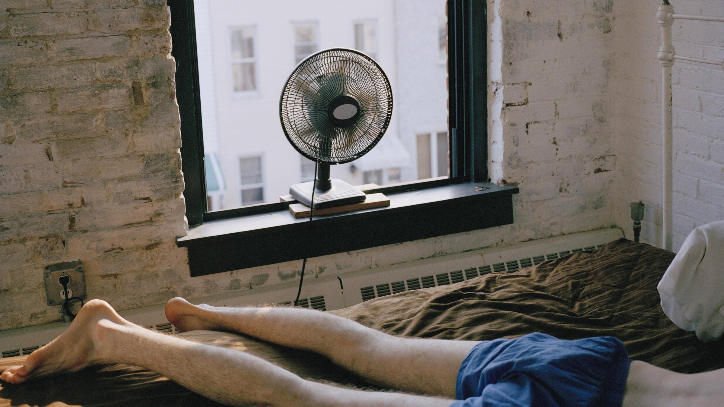 Ein Mann liegt in Unterhose auf einem Bett in einem Zimmer in New York City. Ein Ventilator steht auf dem Fensterbrett.
