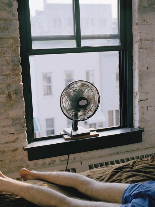 Ein Mann liegt in Unterhose auf einem Bett in einem Zimmer in New York City. Ein Ventilator steht auf dem Fensterbrett.
