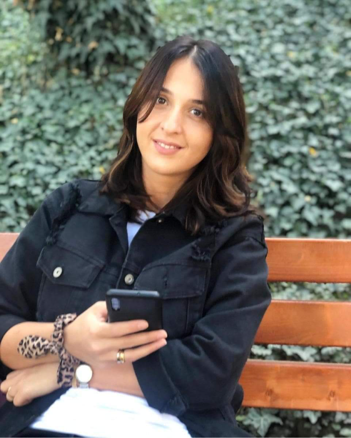 Tamar sitzt auf einer Bank, hat ihr Handy in der hand. Sie hat schwarze, lange Haare und lächelt. Tamar sitzt auf einer Bank, hat ihr Handy in der hand. Sie hat schwarze, lange Haare und lächelt.