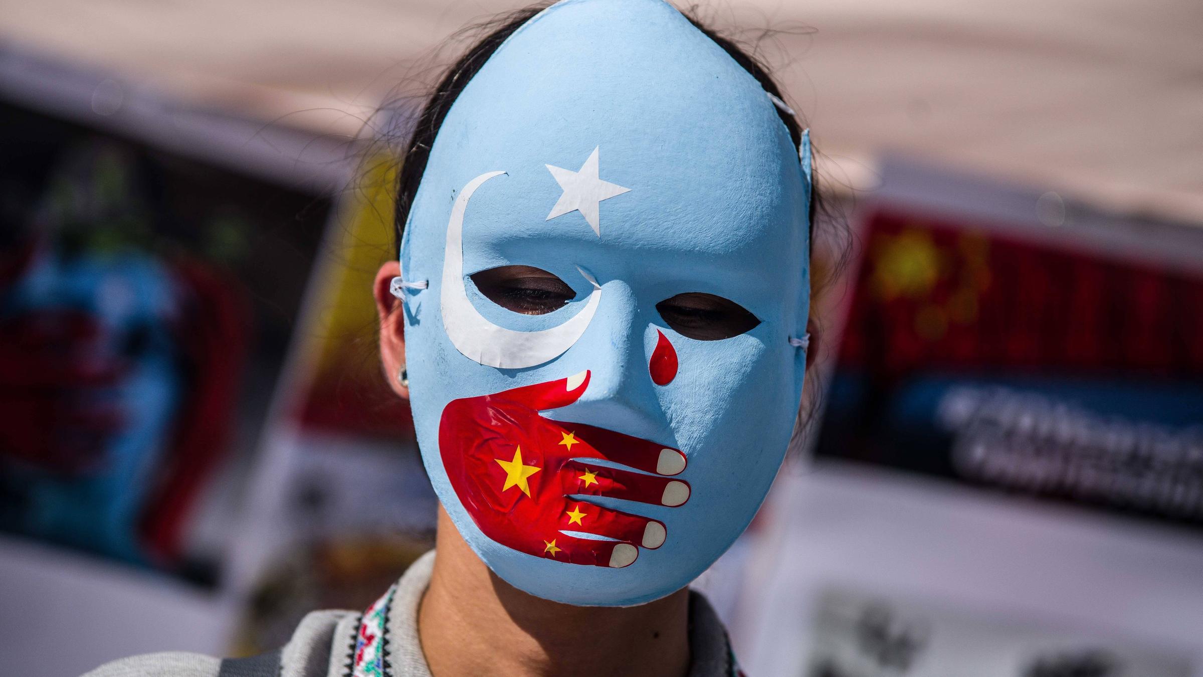 Eine Frau trägt eine blaue Maske mit weißem Stern und weißer Mondsichel. Über den Mund ist eine rote Hand gemalt, die eine chinesische Fahne symbolisiert. Im rechten Augenwinkel ist eine rote Träne. Eine Frau trägt eine blaue Maske mit weißem Stern und weißer Mondsichel. Über den Mund ist eine rote Hand gemalt, die eine chinesische Fahne symbolisiert. Im rechten Augenwinkel ist eine rote Träne.