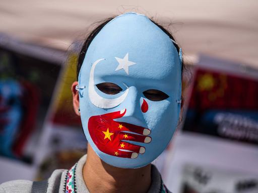 Eine Frau trägt eine blaue Maske mit weißem Stern und weißer Mondsichel. Über den Mund ist eine rote Hand gemalt, die eine chinesische Fahne symbolisiert. Im rechten Augenwinkel ist eine rote Träne. 