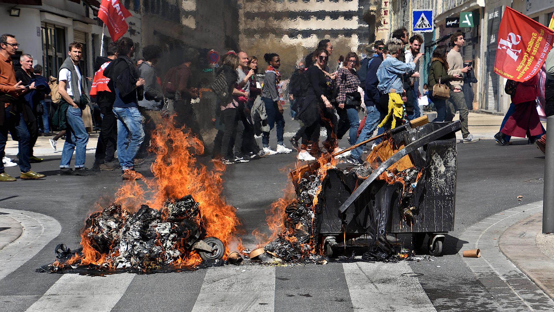 Eine brennende Mülltonne liegt auf der Straße, im Hintergrund ziehen Protestierende vorbei. Eine brennende Mülltonne liegt auf der Straße, im Hintergrund ziehen Protestierende vorbei.