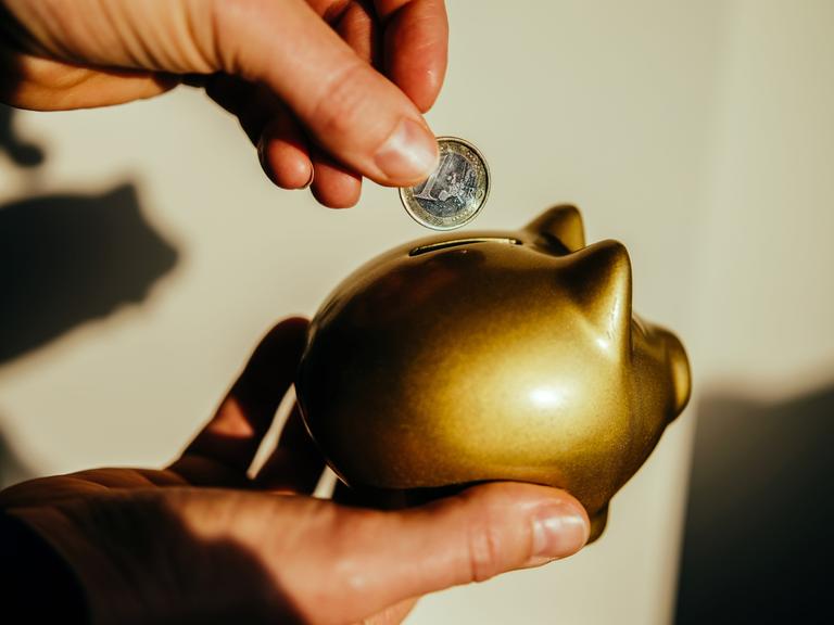 Eine Frauenhand steckt einen Euro in ein goldenes Sparschwein.
