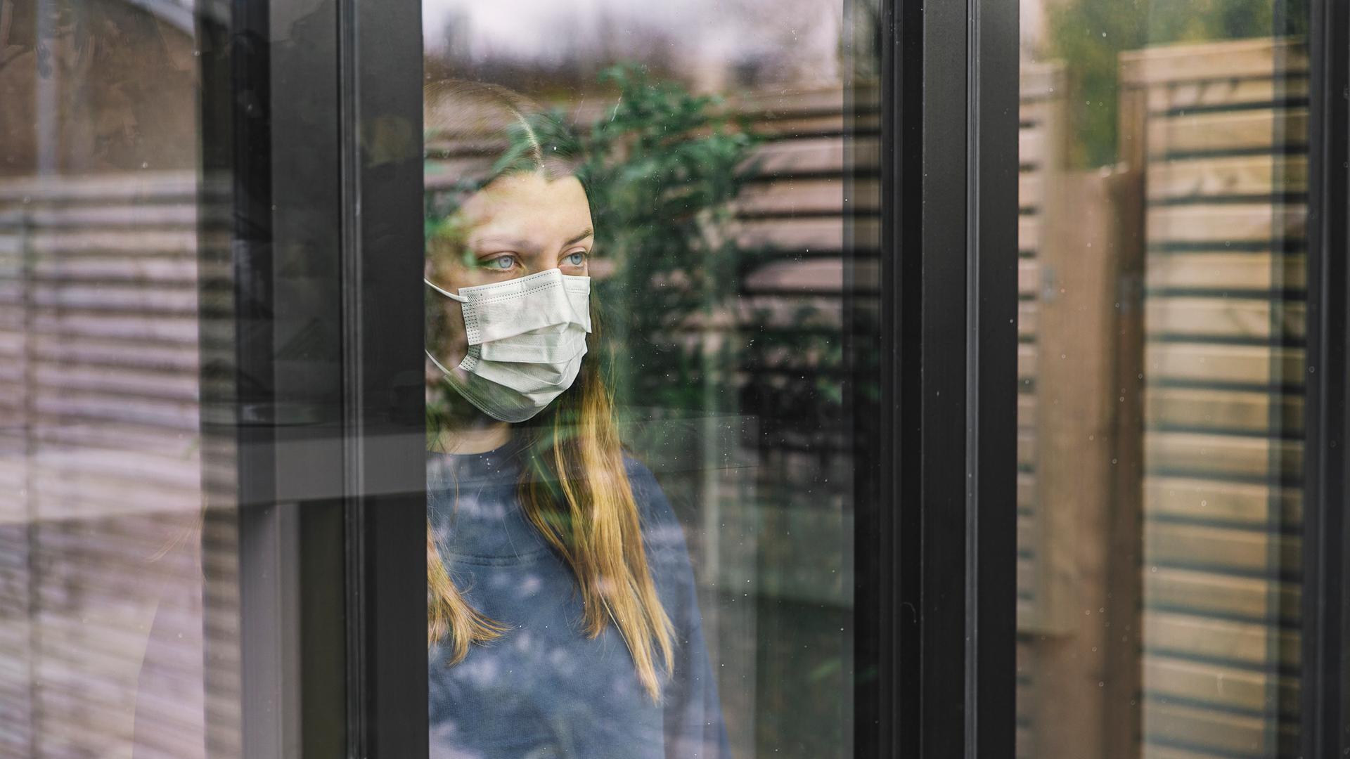 Eine junge Frau mit Maske schaut aus einem Fenster in dem sich ein Zaun und eine Pflanze spiegeln. Eine junge Frau mit Maske schaut aus einem Fenster in dem sich ein Zaun und eine Pflanze spiegeln.