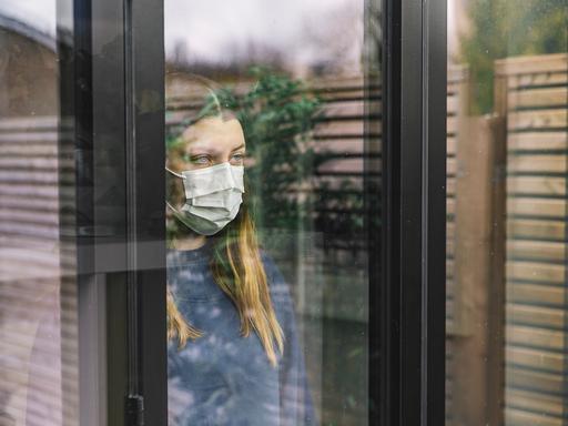 Eine junge Frau mit Maske schaut aus einem Fenster in dem sich ein Zaun und eine Pflanze spiegeln. 