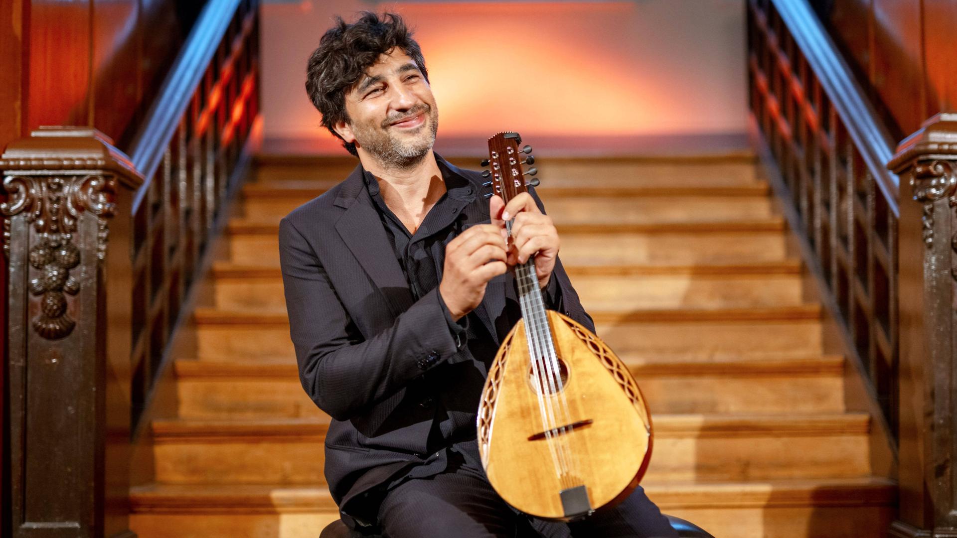 Der Mandolinist Avi Avital sitzt mit seinem Instrument vor einer Holztreppe und lacht in die Kamera.