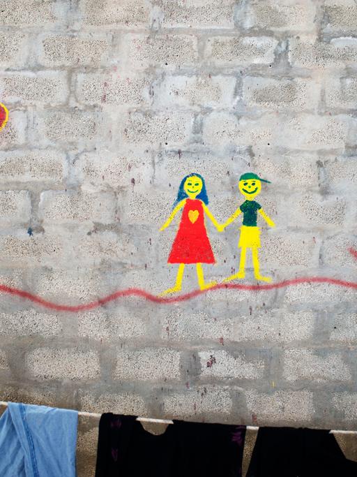 Ein Gemälde auf einer Backsteinwand im Lager Tampakika auf der Insel Chios zeigt ein Mädchen und einen Jungen, die sich an den Händen anfassen. Im Vordergrund hängt Wäsche an einer Leine.