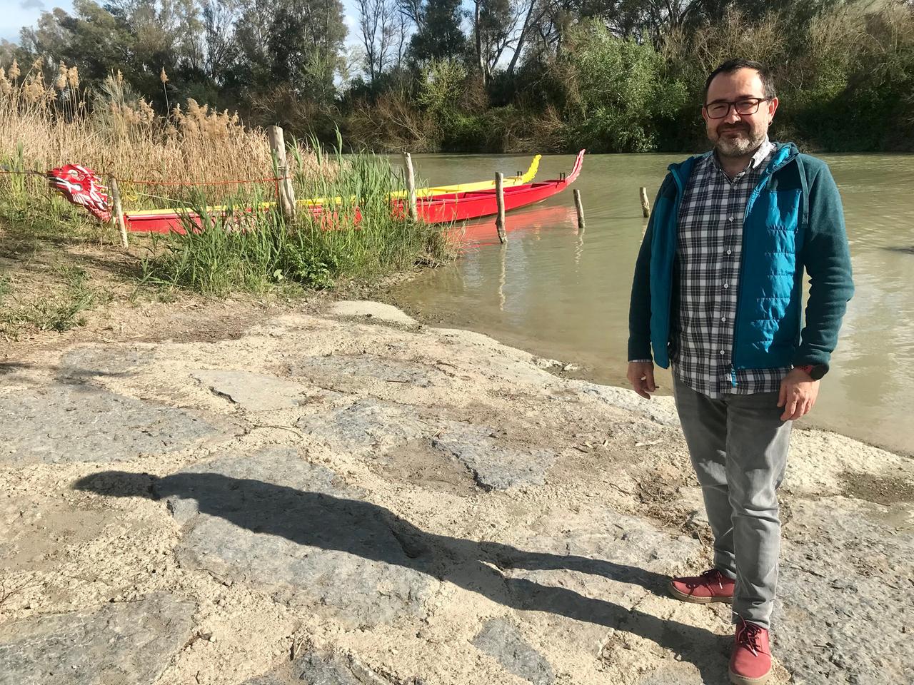 Ein Mann mit dunklem Stoppelbart und Anorak steht am Ufer eines trüben Flusses, an dessen Ufer zwei Kanus liegen.