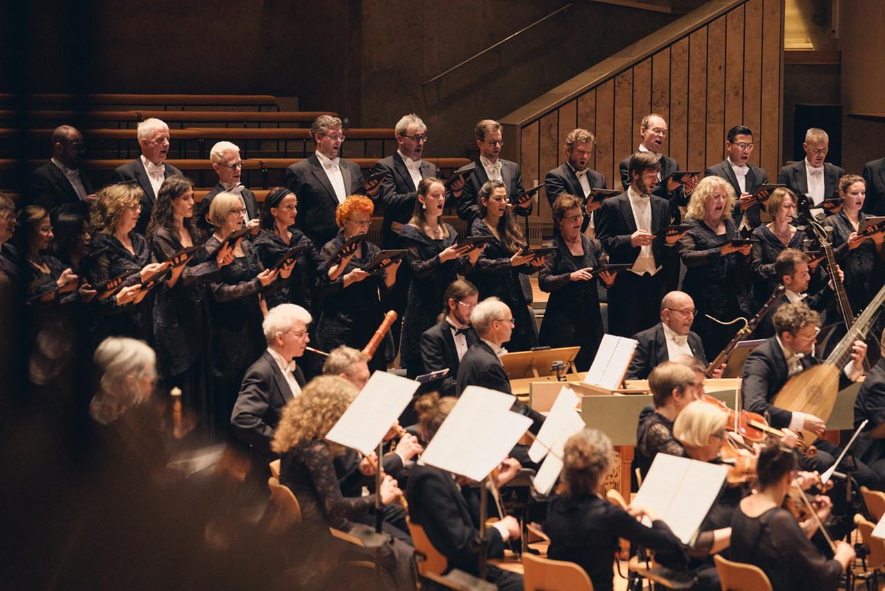 Ein Blick über einer Brüstung hin zur Bühne, auf dem ein Orchester mit historischen Instrumenten sitzt und ein gemischter Chor in festlich-schwarzer Kleidung steht. Ein Blick über einer Brüstung hin zur Bühne, auf dem ein Orchester mit historischen Instrumenten sitzt und ein gemischter Chor in festlich-schwarzer Kleidung steht.