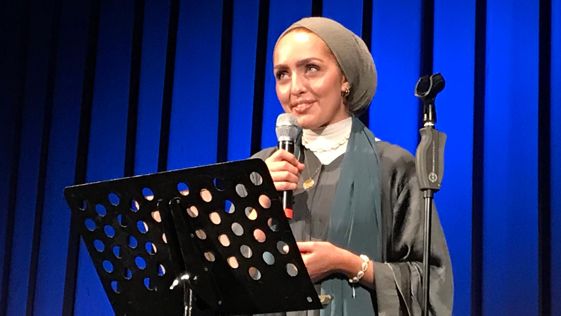 Eine junge Frau im grauen Kostüm, mit Beanie-Mütze spricht in ein Mikrofon und schaut verklärt.