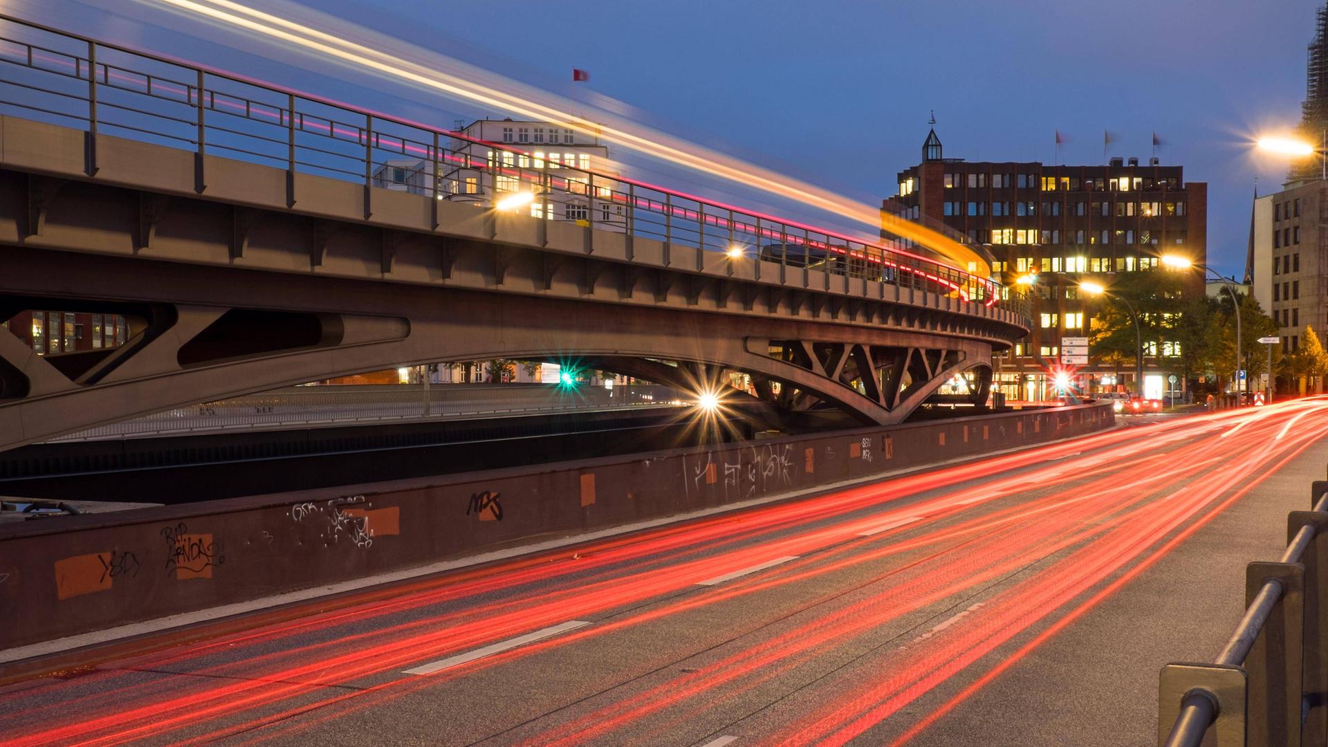 Nacht in einer Stadt: Auf einer von Laternen gesäumten Straße verlaufen leuchtenden roten Linien von fahrenden Autos, links davon eine Brücke mit einer Lichtlinie von einer fahrenden U-Bahn, im Hintergrund ein Haus mit beleuchteten Fenstern. Nacht in einer Stadt: Auf einer von Laternen gesäumten Straße verlaufen leuchtenden roten Linien von fahrenden Autos, links davon eine Brücke mit einer Lichtlinie von einer fahrenden U-Bahn, im Hintergrund ein Haus mit beleuchteten Fenstern.