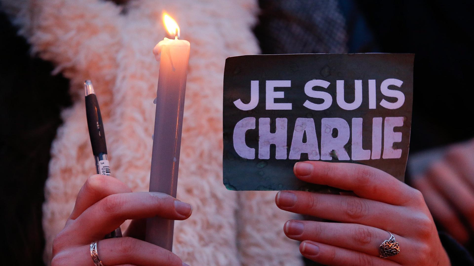 Eine Frau hält eine Kerze, einen Kugelschreiber und eine Schild mit der AUfschrift "Je suis Charlie"