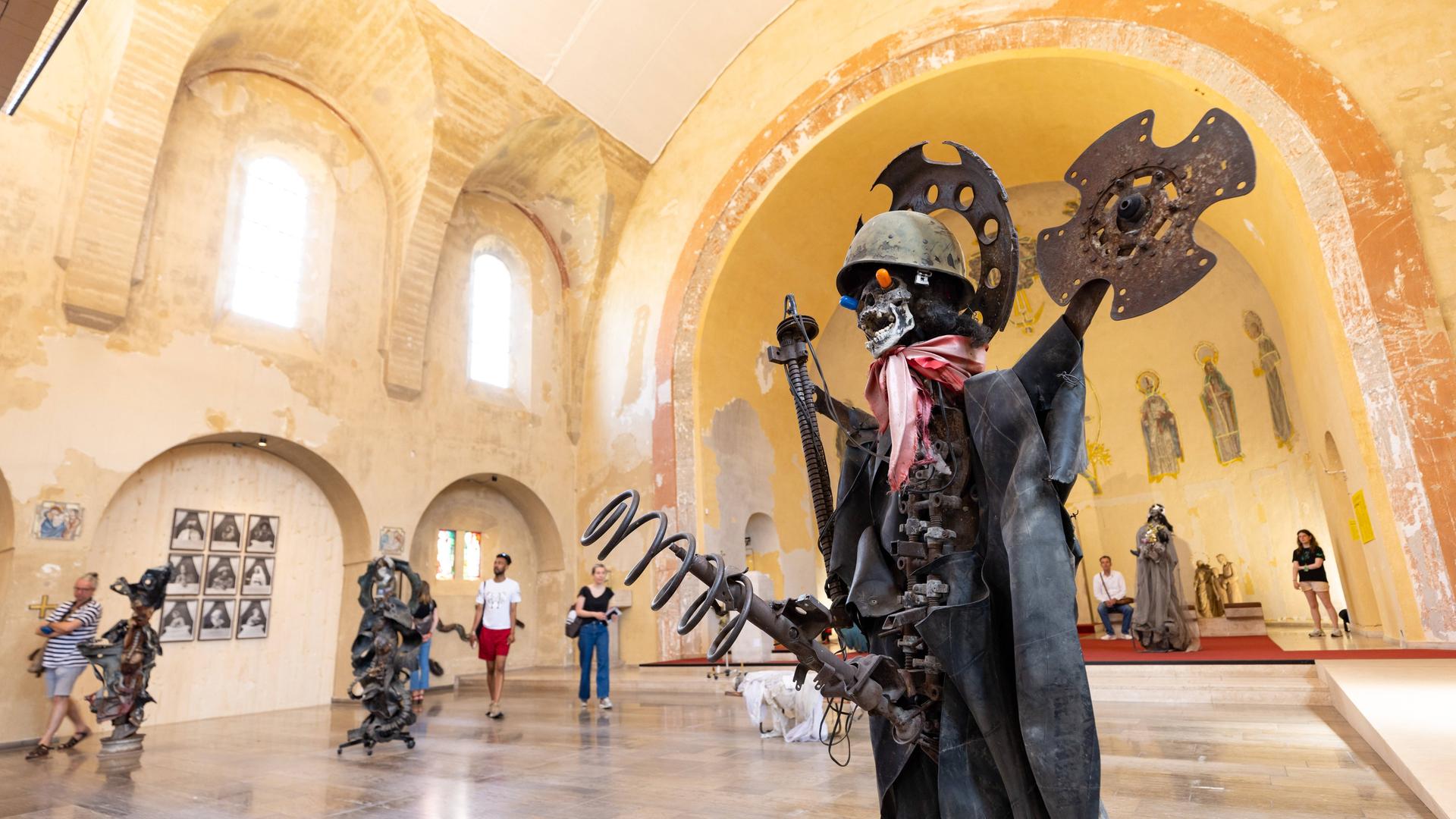 Die aus Maschinenteilen, einem Stahlhelm und Tüchern zusammengesetzte lebensgroße Figur eines Soldaten steht zusammen mit anderen Skulpturen in einer ehemaligen Kirche. Die aus Maschinenteilen, einem Stahlhelm und Tüchern zusammengesetzte lebensgroße Figur eines Soldaten steht zusammen mit anderen Skulpturen in einer ehemaligen Kirche.