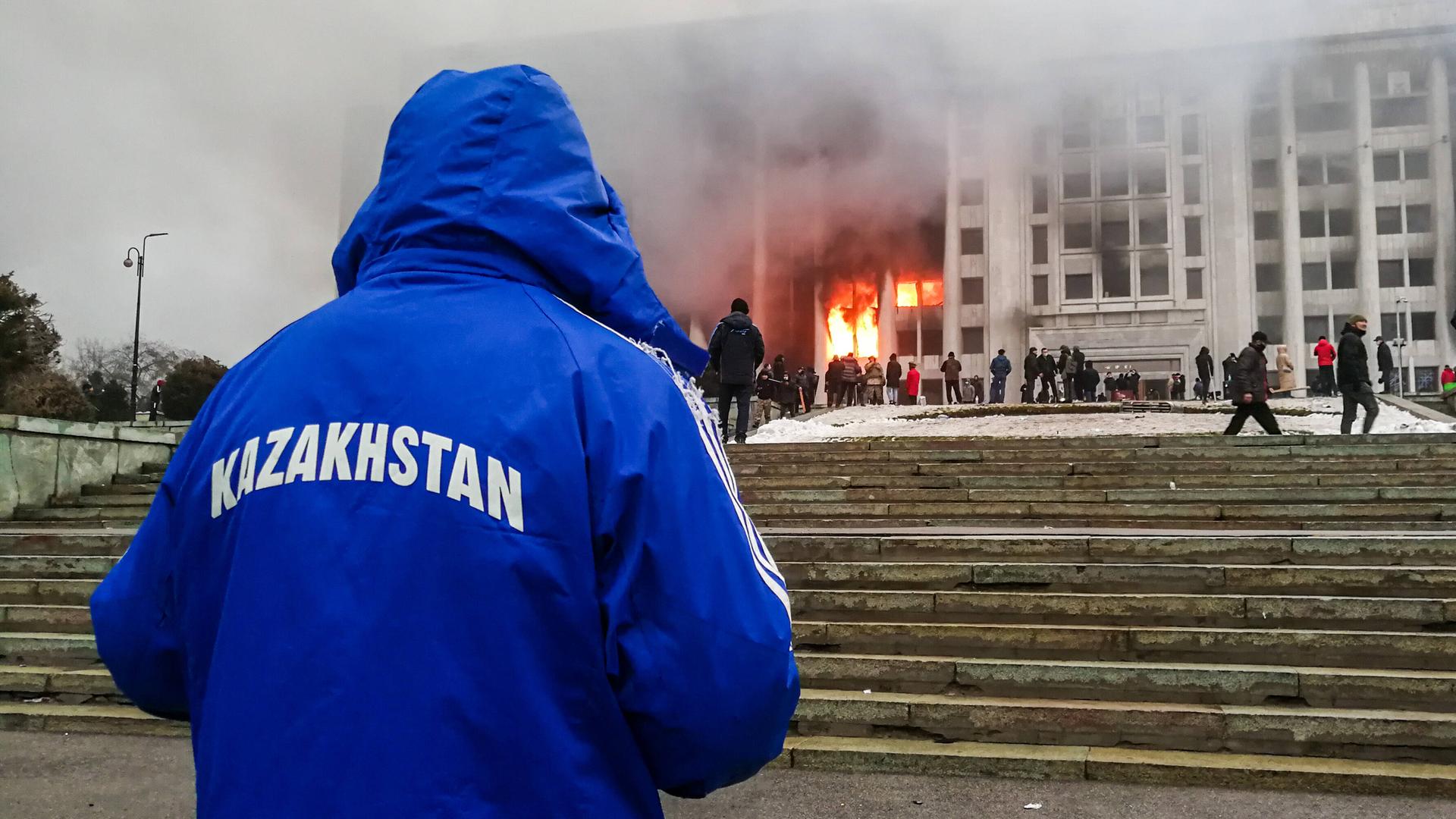 Ein mit dem Rücken zur Kamera stehender Mann beobachtet ein brennendes Regierungsgebäude. Auf der blauen Jacke des Manns steht "Kasachstan" Ein mit dem Rücken zur Kamera stehender Mann beobachtet ein brennendes Regierungsgebäude. Auf der blauen Jacke des Manns steht "Kasachstan"