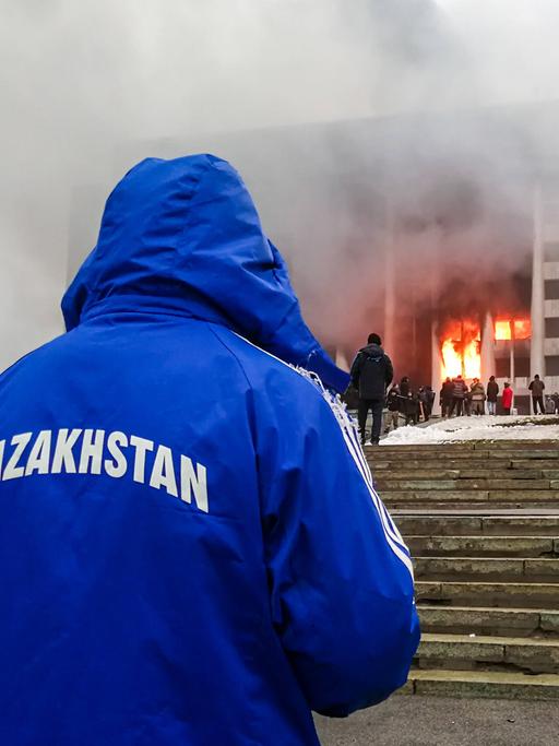 Ein mit dem Rücken zur Kamera stehender Mann beobachtet ein brennendes Regierungsgebäude. Auf der blauen Jacke des Manns steht "Kasachstan"