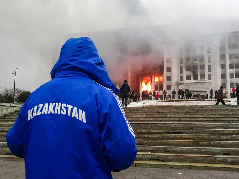 Ein mit dem Rücken zur Kamera stehender Mann beobachtet ein brennendes Regierungsgebäude. Auf der blauen Jacke des Manns steht "Kasachstan"