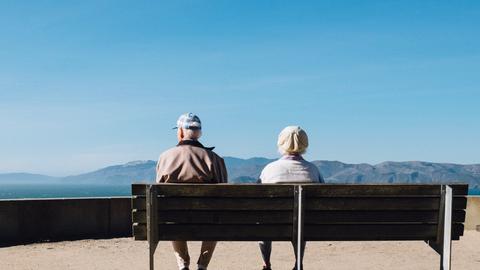 Ein älteres Paar sitzt auf einer Bank und schaut auf eine Berglandschaft. 