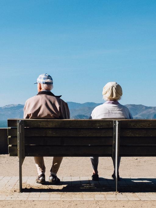 Ein älteres Paar sitzt auf einer Bank und schaut auf eine Berglandschaft. 