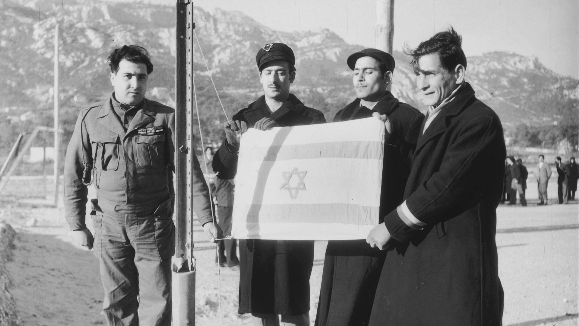 Eine Gruppe von drei jüdischen Männern (Displaced Persons) mit israelischer Fahne vor ihrer Abreise nach Palästina neben einem Soldaten (um 1948). Eine Gruppe von drei jüdischen Männern (Displaced Persons) mit israelischer Fahne vor ihrer Abreise nach Palästina neben einem Soldaten (um 1948).