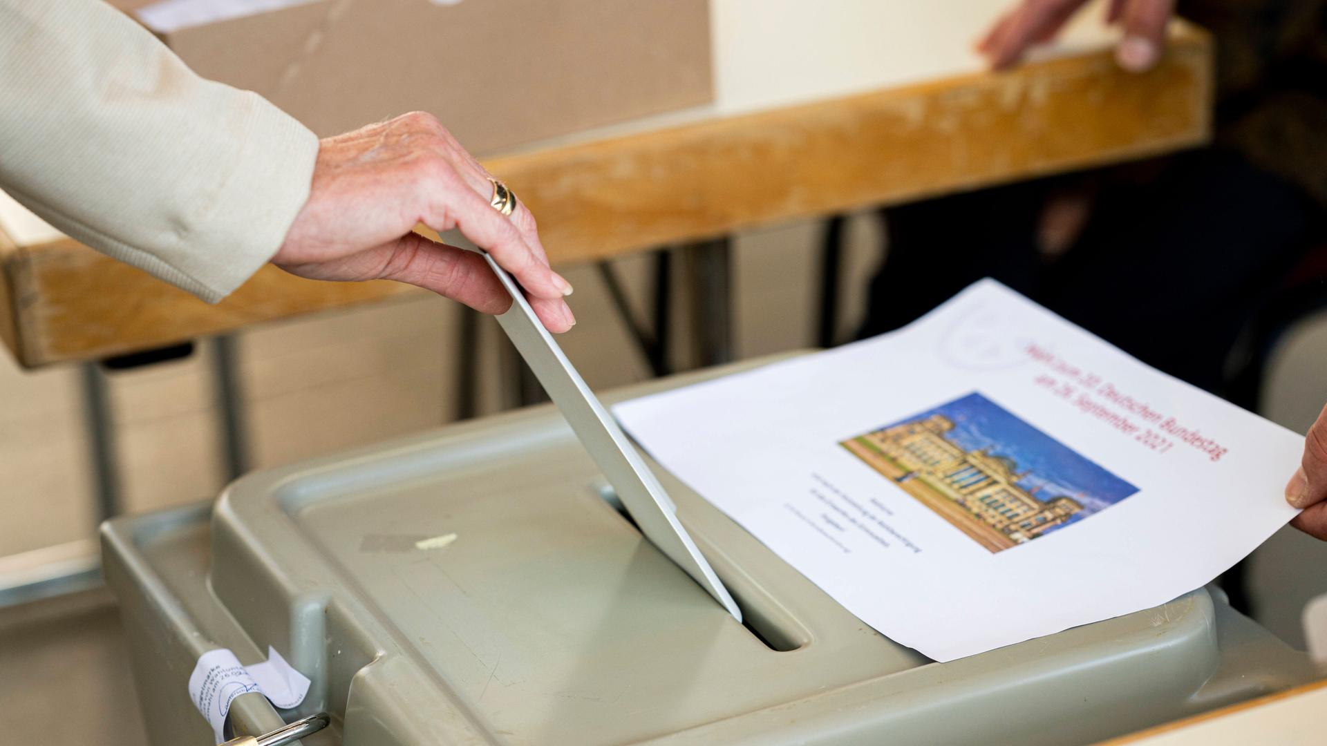 Zu sehen ist die Hand einer Frau in einem Wahllokal , die ihren Stimmzettel in den Schlitz einer Wahlurne steckt.