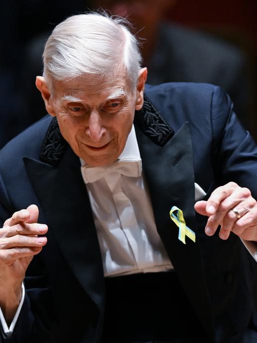 Ein Herr von 95 Jahren hält die Hände vor sich, er gestikuliert, dirigiert ein Orchester. Er blickt aufmerksam und vergnüngt. Er trägt einen dunkelblauen Anzug.